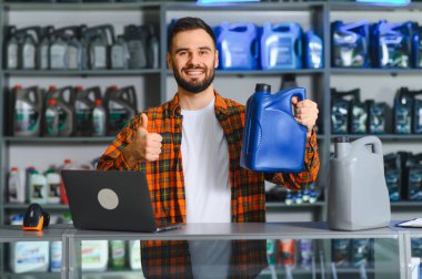 Motor yağı şişesi tutan sakallı mutlu bir adam, bir tamirhanede dizüstü bilgisayarla tezgahın arkasında dikiliyor.