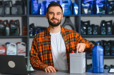 Tamircide elinde araba aküsü ve yağ şişesiyle flanel giymiş gülümseyen tamirci parçalarını ve bakım tavsiyelerini sunuyor.