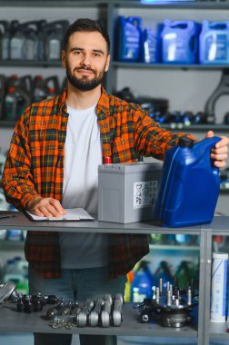 Tamirci bir oto yedek parça dükkanında elinde bir motor yağı kabı tutuyor ve satılık bir araba aküsü sergiliyor.