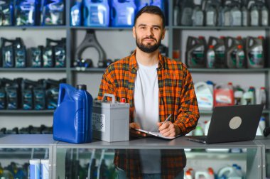 Tamirci panoya yazı yazıyor. Motor yağı ve araba aküsüyle tezgahta duruyor. Raflar arkasında duruyor.