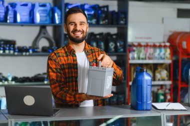 Otomobil mağazasında çalışan, elinde araba aküsü olan, arkasında kayganlaştırıcı ve otomotiv ürünleri olan bir adam.