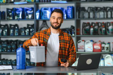 Gülümseyen ve elinde araba aküsü tutan bir işçi, bir oto yedek parça dükkanının tezgahında duruyor ve raflarında motor yağı var.
