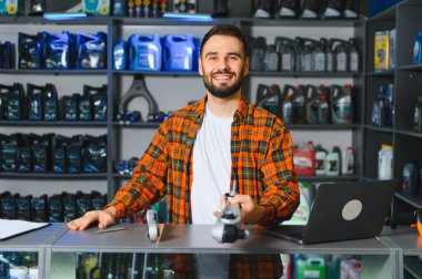 Bir otomotiv malzemeleri dükkanında tezgahın arkasında duran genç bir adam, elinde bir araba parçası, kameraya bakıyor.