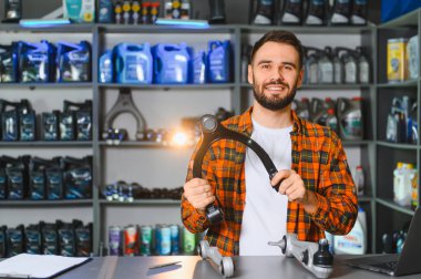 Askı kontrol kolu tutan genç bir adam, oto yedek parça mağazasında gülümsüyor. Kayganlaştırıcı ve yedek parçalarla dolu raflar