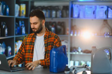 Genç bir erkek çalışan oto yedek parça mağazasının tezgahında dizüstü bilgisayar kullanıyor. Çevresinde motor yağı ve araba parçaları var.