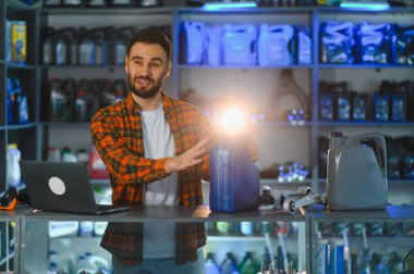 Adam konuşuyor, elinde motor yağı kabı var, yedek parça dükkanında duruyor. Dizüstü bilgisayar ve araba parçaları tezgahın üzerinde.