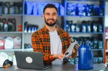 Tezgahın arkasında duran gülümseyen adam, elinde süspansiyon parçası ve arka planda araba ürünleri rafları olan bir yağ kabı tutuyor.