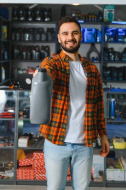 Ekose gömlekli gülümseyen adam otomotiv parçaları mağazasının içindeki kameraya doğru motor yağı şişesi tutuyor, kendinden emin satış teknisyeni