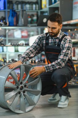 İş tulumlu tamirci ve ekose gömlekli bir otomobil alaşımlı tekerleği inceliyor, lastik dükkanındaki çatlakları ya da deformasyonları kontrol ediyor.