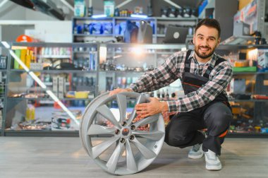 Gülümseyen araba tamircisi yeni bir alaşım tekerleği tutuyor, bir araba parçacıda duruyor, araç servisine ya da tamirine hazır.