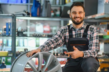Yeni bir alaşım tekerleği tutan otomobil parçaları işçisi, özel bir araba tamirhanesinde kameraya bakıp gülümsüyor.