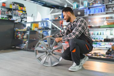 Bir otomotiv dükkanında çalışan bir adam, modern bir araba jantı tutuyor ve rafların önünde ürünlerle dolu duruyor.