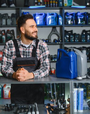 Oto yedek parça mağazasında duran erkek işçi, başka tarafa bakarken gülümsüyor. Arkasında motor yağı ve araba parçaları rafları var.