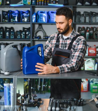 Üniformalı, kendine güvenen sakallı tamirci elinde motor yağı şişesiyle otomotiv sıvılarıyla dolu rafların önünde duruyor.