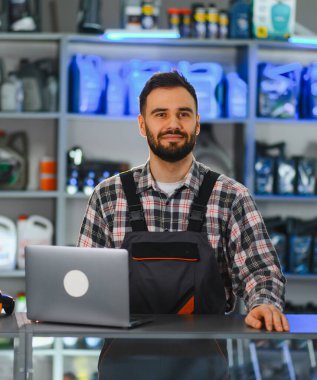 Oto yedek parça mağazasının önünde dizüstü bilgisayar kullanan ve etrafı araba ürünleriyle çevrili mutlu bir şekilde gülümseyen bir erkek işçi.