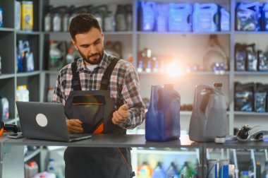 Oto yedek parça mağazası çalışanı üniforma giyiyor dizüstü bilgisayara bakıyor, ürünleri seçiyor, tezgahında yağ şişeleri ve rafları var.