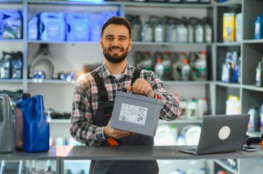 Gülümseyen tamirci oto yedek parça mağazasında araba aküsünü sunuyor. Profesyonel otomotiv hizmeti ve güvenilir ürünler sağlanıyor