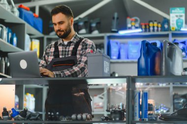 Bir oto yedek parça mağazasında envanter yönetiminde dizüstü bilgisayar kullanan işçi. Kutuları ve araba aküsü olan raflar.