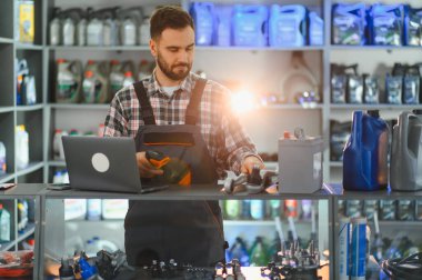Envanter yönetimi için barkod tarayıcı ve laptop kullanan otomobil parçaları mağaza çalışanı, araba aküsü ve yağ ile tezgahta duruyor