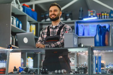 Araba aküleri ve yağ bidonlarıyla çevrili bir oto yedek parça dükkanında dizüstü bilgisayarda çalışan gülümseyen bir tamirci.