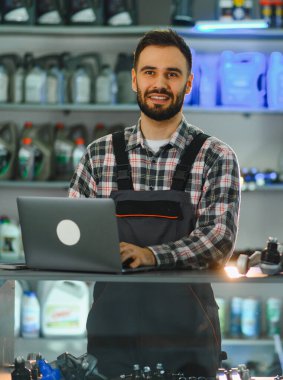 Tamirci üniformalı mutlu sakallı adam gülümsüyor. Bir otomotiv dükkanında dizüstü bilgisayarını envanter veya müşteri siparişi olarak kullanıyor.