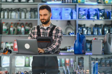 Otomobil parçaları mağazasında tezgahta çalışan perakende asistanı, dizüstü bilgisayarda yazıyor, motor yağı ve araba aküsü sergileniyor