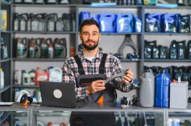 Erkek tamirci, bir araba için yeni bir süspansiyon parçası gösteriyor. Tezgahında yağlar, dizüstü bilgisayarı ve arkasında da ürünler var.