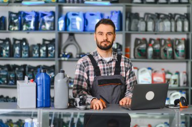 Oto yedek parça işçisi dizüstü bilgisayarı, araba aküsü ve yağıyla tezgahın arkasında duruyor, envanter ve satışları yönetiyor.