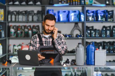 Erkek tamirci telefonla konuşuyor, pano tutuyor ve bir araba tamirhanesinde dizüstü bilgisayar kullanıyor.
