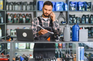 Otomobil yedek parça işçisi süspansiyon parçası tutuyor, dizüstü bilgisayar kullanıyor ve bir araba servis merkezinde cep telefonuyla konuşuyor.