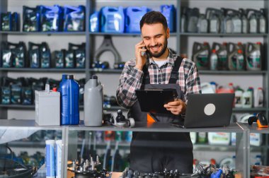 Bir otomotiv parçacıda çalışan gülümseyen erkek çalışan, cep telefonuyla konuşan, dosyadaki siparişleri kontrol eden.