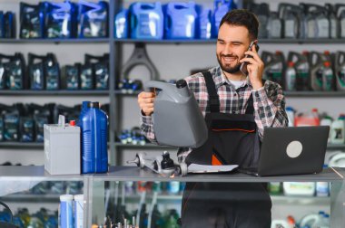 Otomobil servisi teknisyeni telefonda araba parçaları ve yağ hakkında konuşuyor. Atölye tezgahında dizüstü bilgisayar, pil ve sıvı şişelerle çalışıyor.