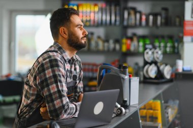 Ekose gömlekli ve tulumlu genç sakallı tamirci servis gişesinde dizüstü bilgisayarı, motor yağı ve araba parçaları ile işe odaklanmış.