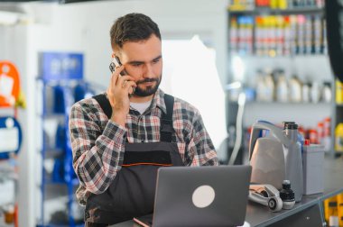Tamirci cep telefonuyla konuşuyor ve yedek parça ve yağlarla çevrili bir araba servis istasyonunda dizüstü bilgisayarla çalışıyor.