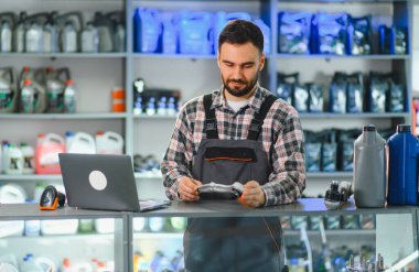 Tulumlu ve ekose gömlekli bir erkek işçi bir araba servisinde ve parça dükkanında bilgisayar masasında bir otomotiv parçasını tarıyor.