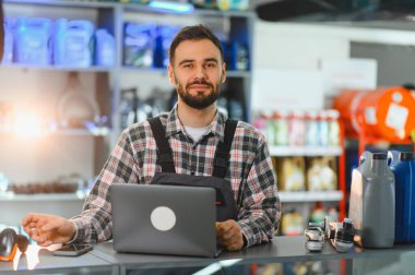 Tulumlu ve ekose gömlekli genç adam gülümsüyor, oto yedek parça mağazasında dizüstü bilgisayar kullanıyor.