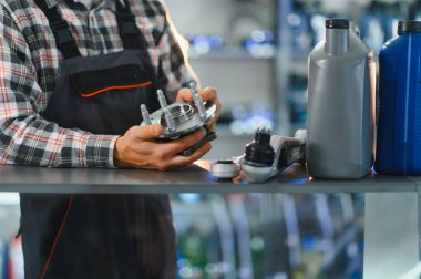 Tamircide yeni tekerlek merkezi montajını otomobil tamircisi denetliyor. Motor yağı şişeleri ve otomobil parçaları rafta.