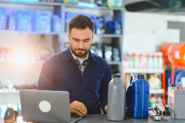Bir otomotiv hizmet merkezinde dizüstü bilgisayar ve satış yapan erkek işçi, tezgahında motor yağı ve araba aküsü bulunuyor.