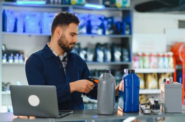 Bir oto yedek parça mağazasında motor yağını tarayan erkek işçi, envanteri dizüstü bilgisayar ve barkod tarayıcı ile yönetiyor.