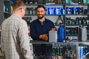 Gülümseyen tamirci araba aküsü ve motor yağı satan bir mağazada erkek müşteriye yardım ediyor.