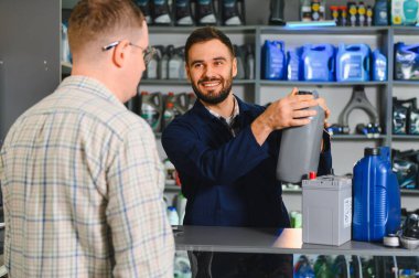 Oto yedek parça mağazası çalışanı müşteriye yardım ediyor, elinde bir şişe motor yağı tutuyor. Araba aküsü ve daha fazla yağ şişesi raflarda.