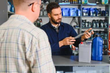 Oto yedek parça işçisi dükkandaki bir müşteriye araba parçalarını açıklıyor. Süspansiyon kolu, araba aküsü ve motor yağı gösteriliyor