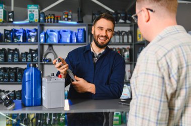 Üniformalı otomobil işçisi araba parçası tutuyor, otomotiv ürünleriyle endüstriyel rafların önünde müşteriyle konuşuyor.