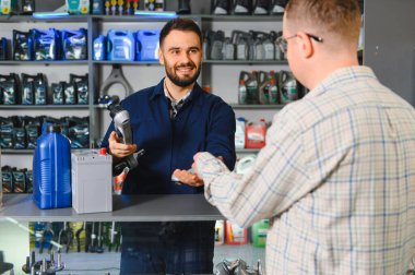 Satıcı gülümsüyor, araba parçası tutuyor, araç bakım malzemeleri ve aksesuarları seçen müşteriye yardım ediyor.