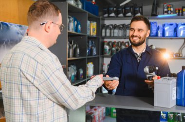 Araba yedek parça satıcısı müşteriye yardım ediyor, araba parçası tutuyor, tezgahta akü ve yağ şişeleri var.