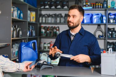 Bir süspansiyon parçası tutan oto yedek parça satıcısı, mağaza tezgahında akıllı telefon ile temas kurmadan ödeme yapan müşteri ile etkileşim