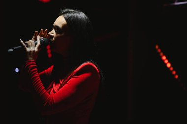 Kadın sanatçı yoğun bir şekilde mikrofona şarkı söylüyor, karanlık bir sahnede spot ışıklarıyla aydınlanıyor bulanık ışıklarla.