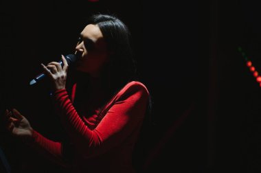 Kırmızı elbiseli bir kadın performans sırasında mikrofona şarkı söylüyor, karanlık bir sahnede spot ışıklarıyla aydınlanıyor.