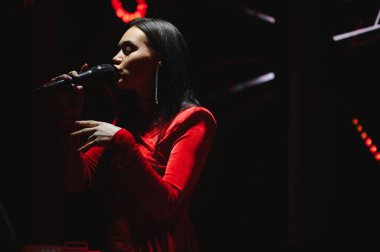Kırmızı elbiseli genç bir kadın mikrofon tutarak şarkı söylüyor, kırmızı spot ışıklarıyla karanlık bir sahnede müzik yapıyor.