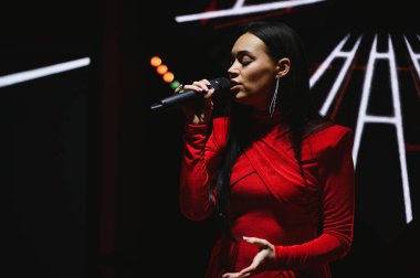 Kırmızı elbiseli genç bir kadın sahne ışıkları altında mikrofona şarkı söylüyor. Kendine güvenen, göz alıcı bir şekilde canlı müzik yapıyor.